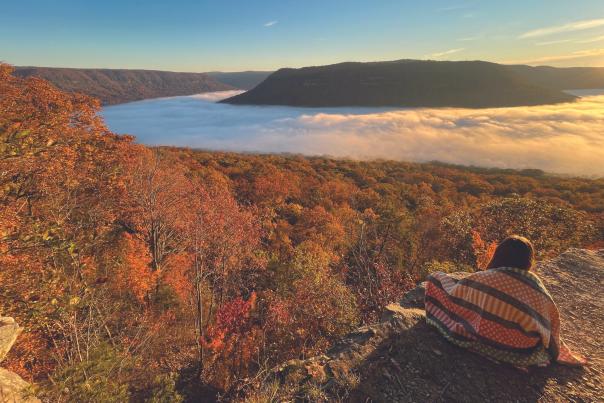 woman sits at Snooper's Rock wrapped in quilt watching sunrise at overlook
